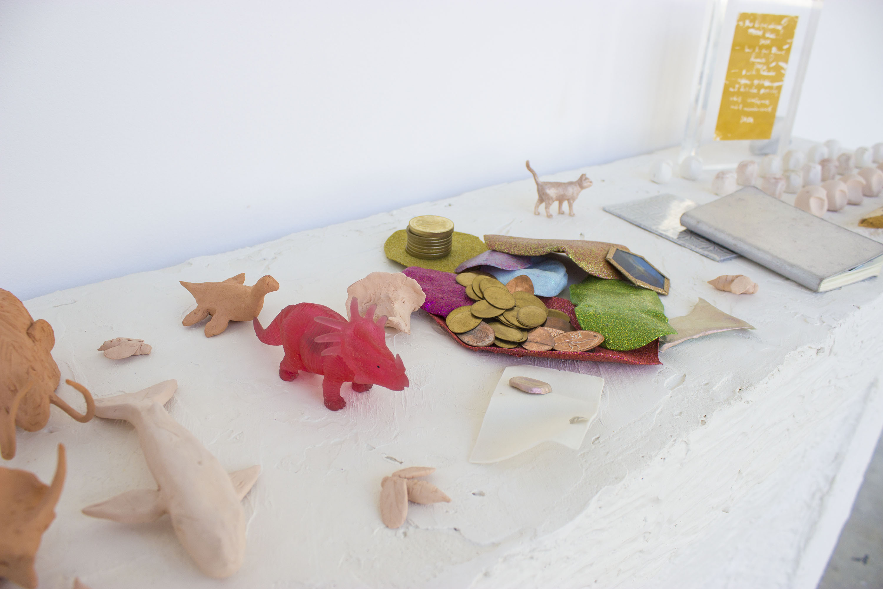 Gros plan sur les choses posées sur la cheminée : des mammouths en céramique rouge, un cachalot en cérmaique rose pâle, un plésiosaure en céramique rouge, 3 abeilles en céramique rose pâle, un tricératops en plastique transparent rouge, des slime séchés sur lesquels sont posés des pièces ovales en métal, une figurine de chat métallisée, des bibelots de chouette en céramique, des carnets et une sérigraphie dorée d'un texte écrit à la main dans un cabre en plexiglas transparent